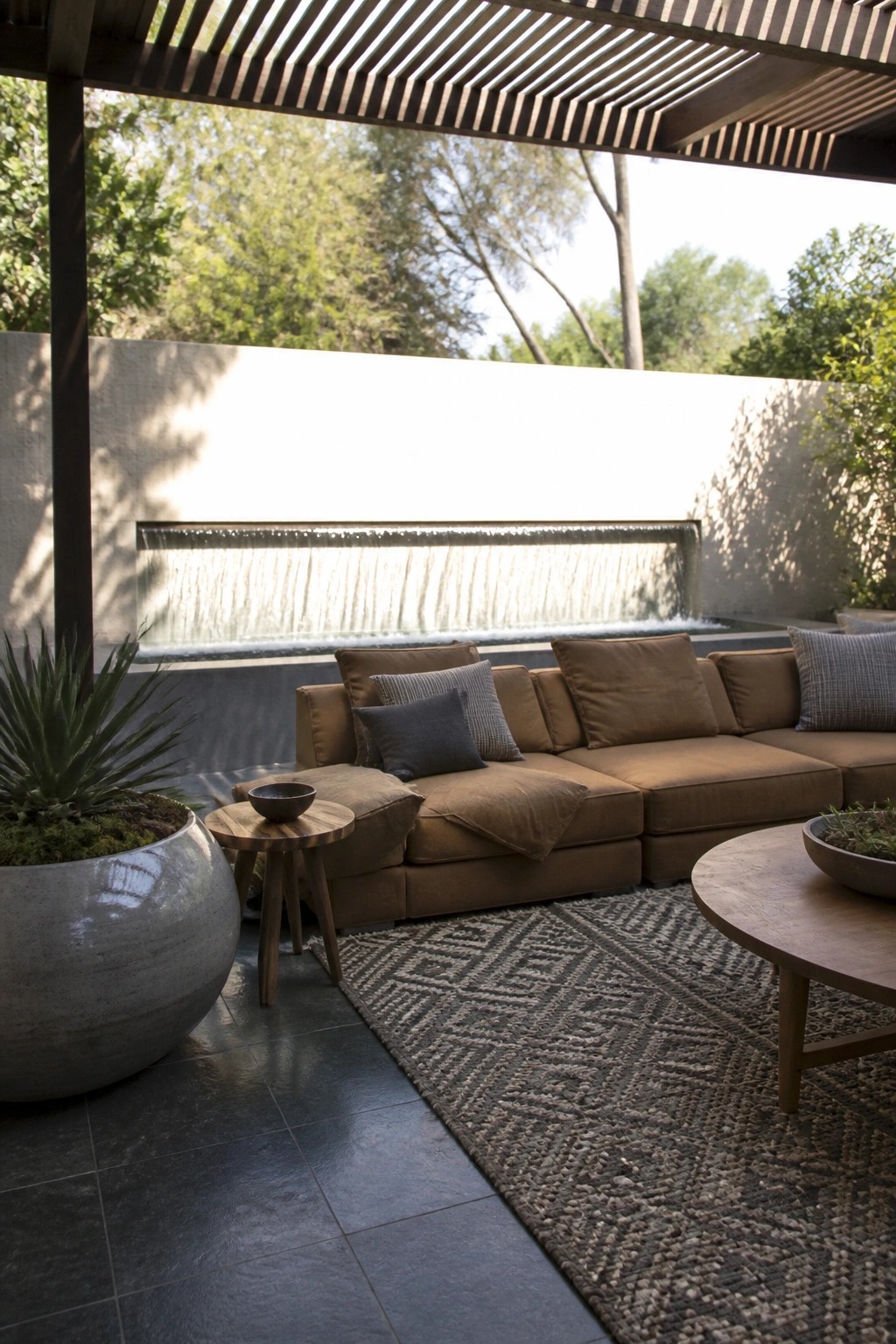A large brown sectional sofa with deep cushions sits on a patterned rug under a wooden pergola, facing a long water wall in an outdoor patio area.