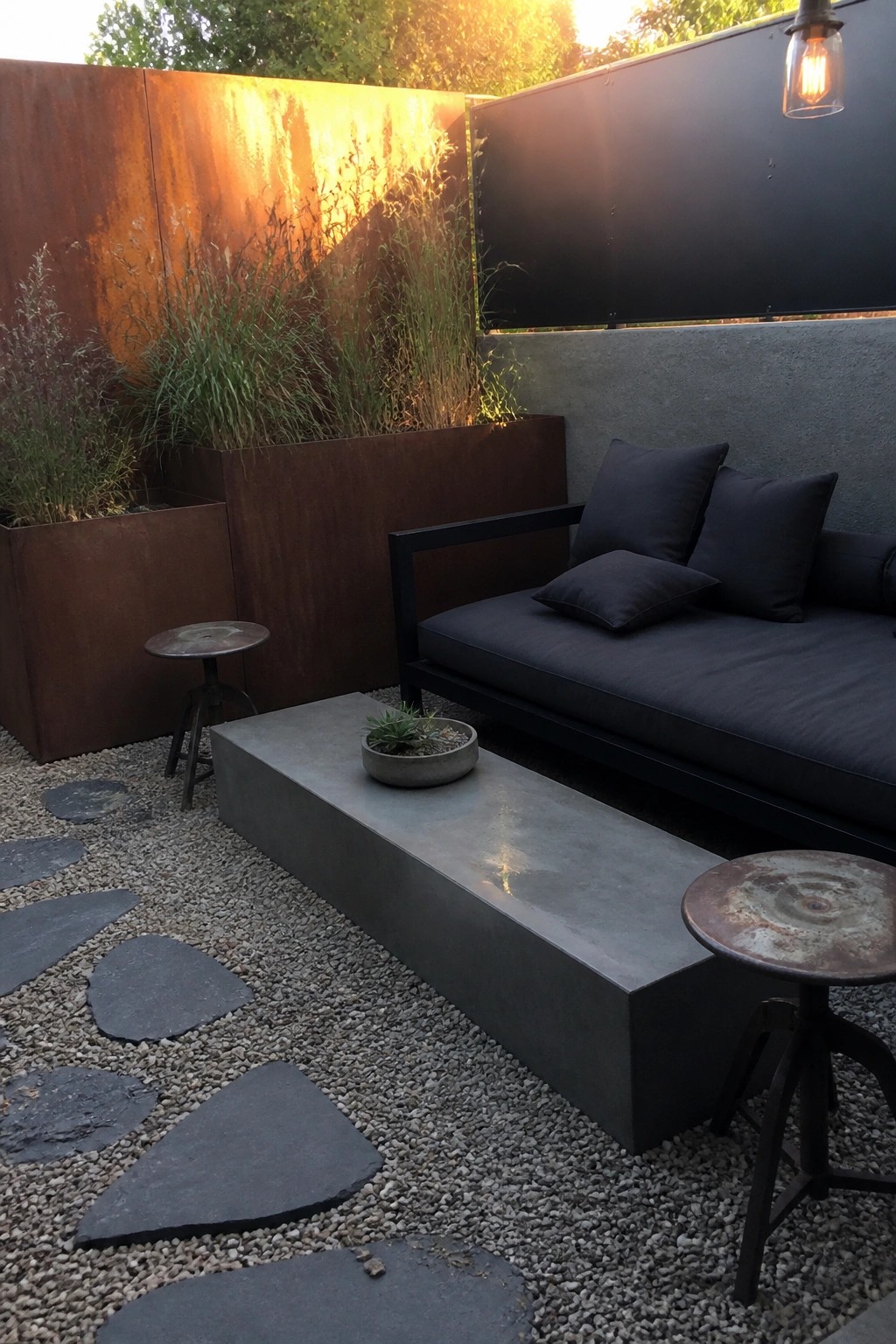 A patio with a long dark lounge sofa with cushions, a concrete bench holding a small plant, gravel ground with stepping stones, metal planters with tall grasses, and two round metal stools.