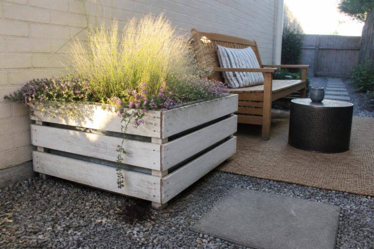 19 Creative DIY Pallet Planter Ideas Built Around Simple Box Designs A weathered white pallet planter box with grasses and pink flowers sits on gravel beside concrete pavers and a wooden bench on an outdoor patio.