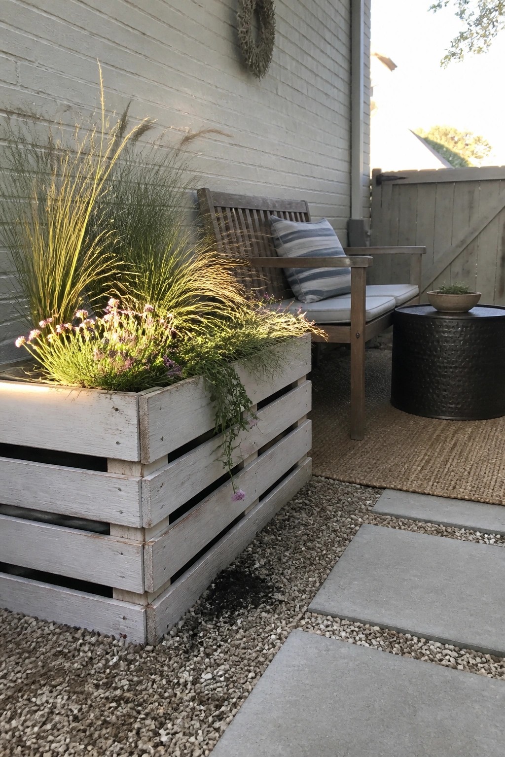 A weathered white pallet planter box with grasses and pink flowers sits on gravel beside concrete pavers and a wooden bench on an outdoor patio.