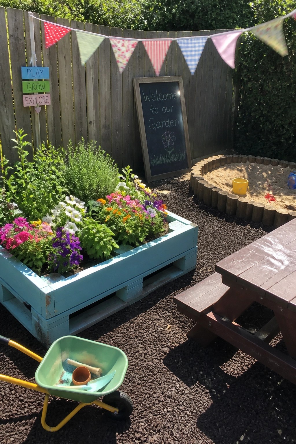 A blue painted wooden pallet planter box filled with colorful flowers sits in a backyard garden beside a sandpit and picnic table.