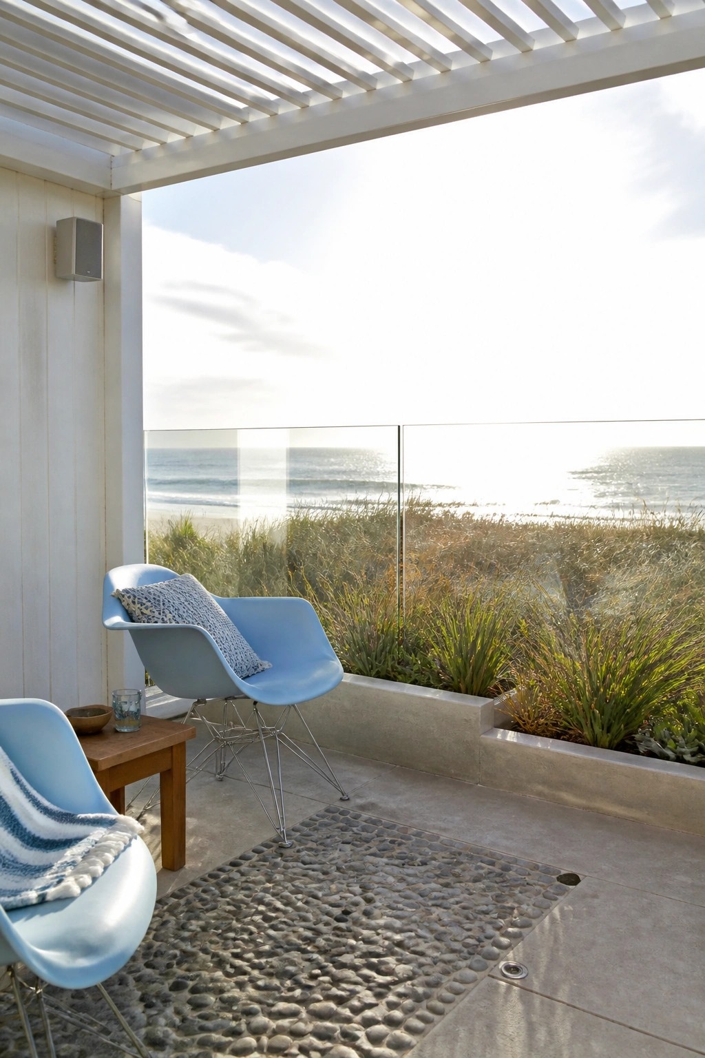 Outdoor patio with light blue molded chairs and a small wooden table under a white slatted pergola, next to a glass railing with an ocean view.