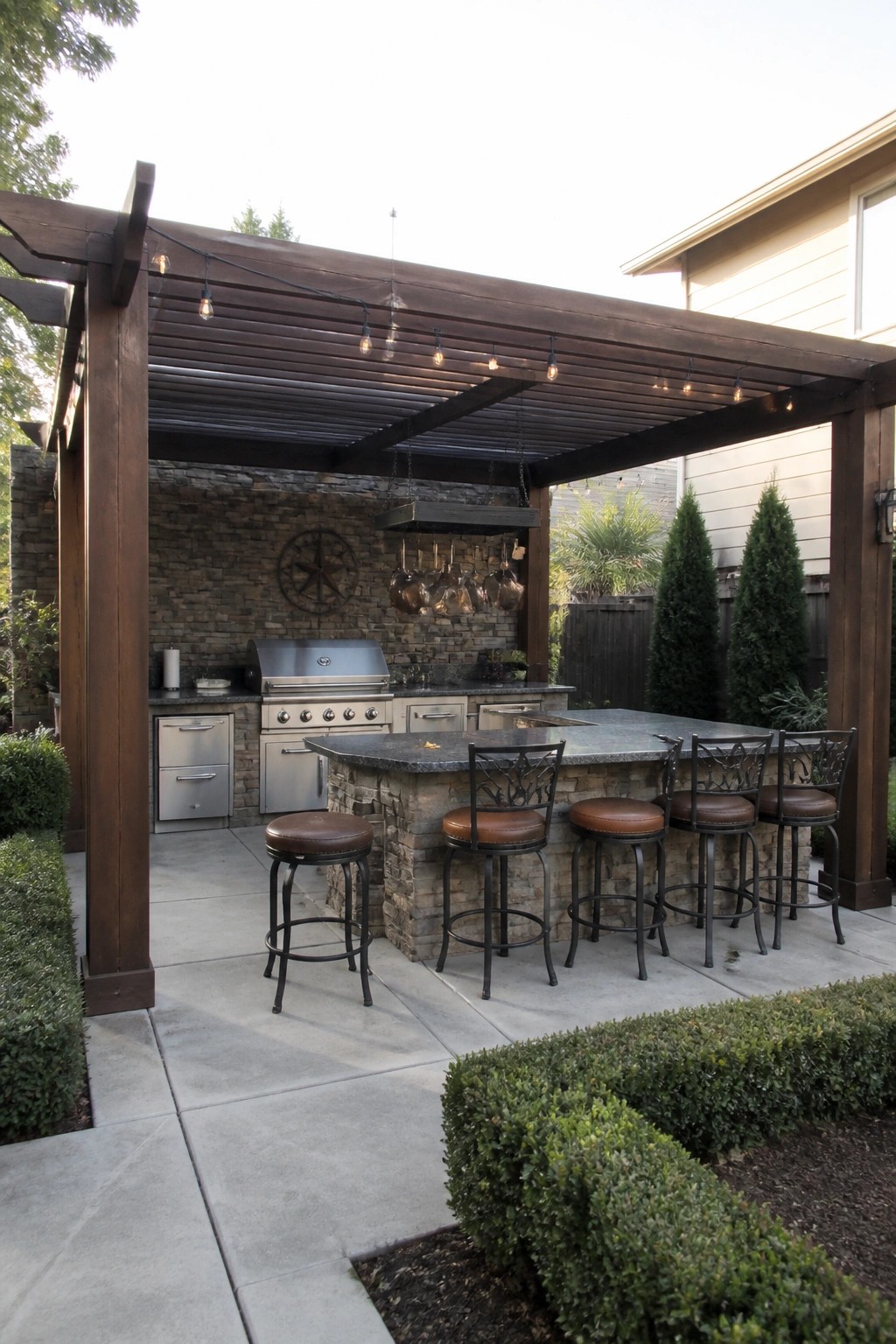 An outdoor patio under a dark wooden pergola with a slatted roof, showing a stone kitchen island, bar stools, grill, and hanging string lights.