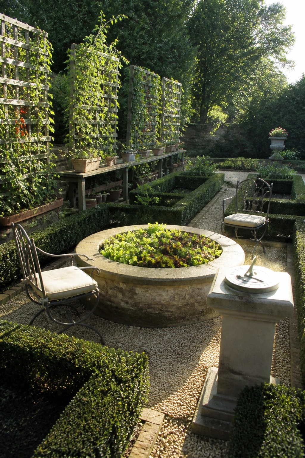 Circular stone planter of leafy greens with chairs, sundial, trellises, and hedges.