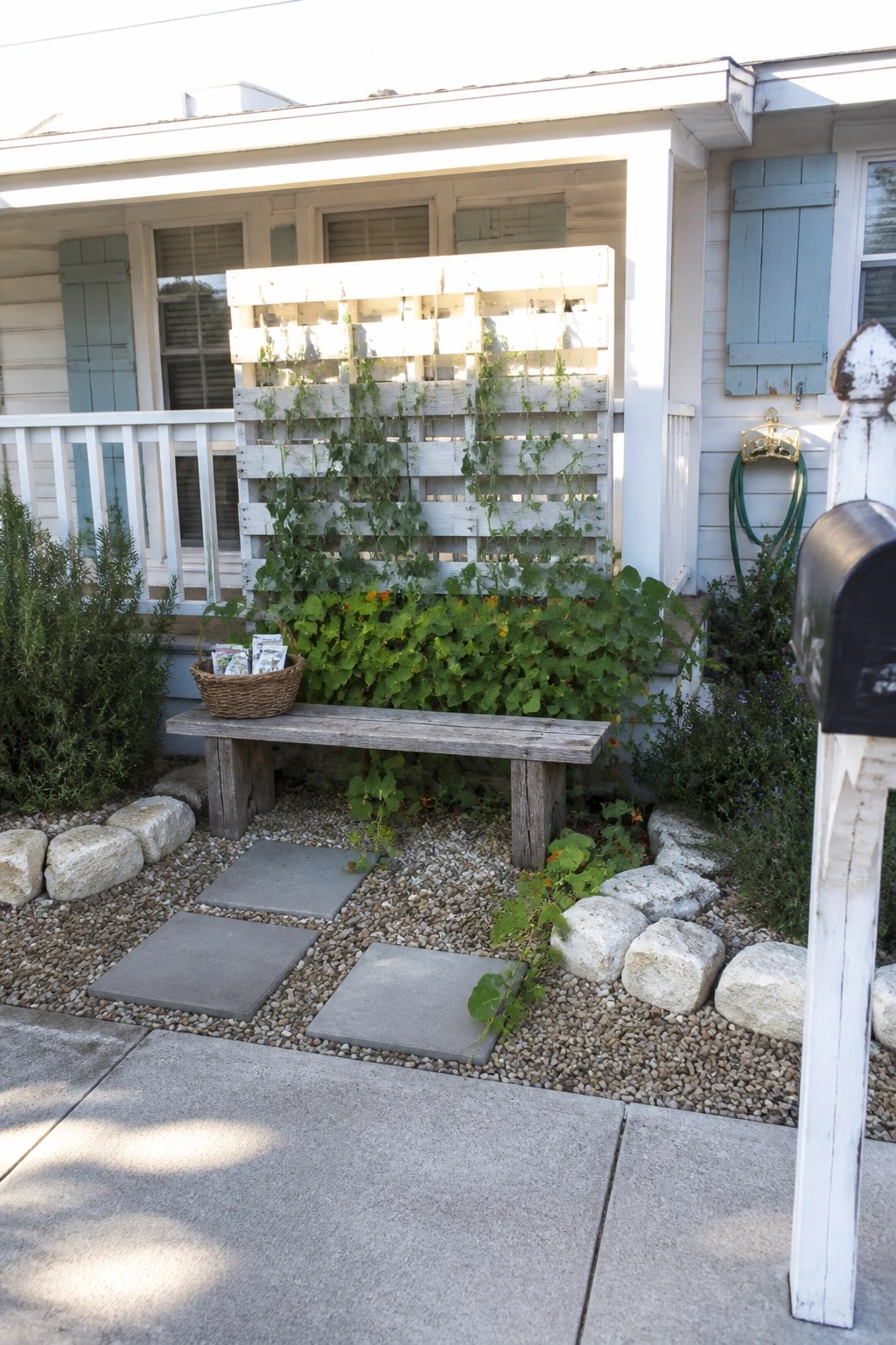 A wooden pallet mounted on the side of a house with green climbing vines, a wooden bench in front, and gravel with stepping stones leading to the porch.