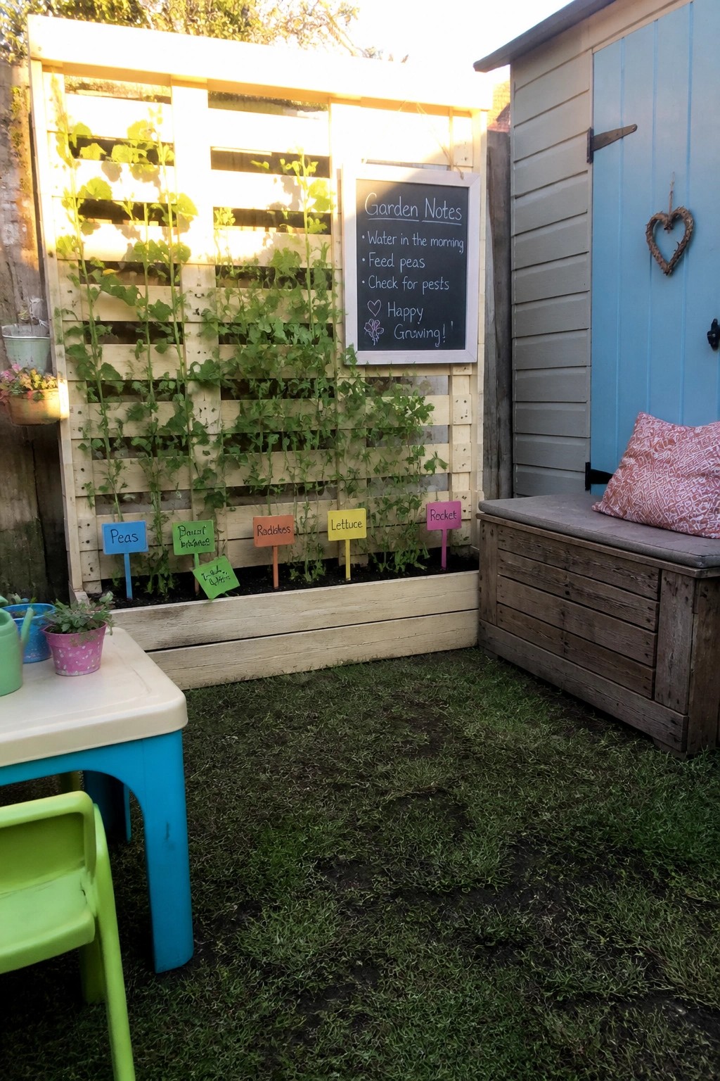 Vertical pallet garden with labeled plants, chalkboard notes, blue shed and wooden bench