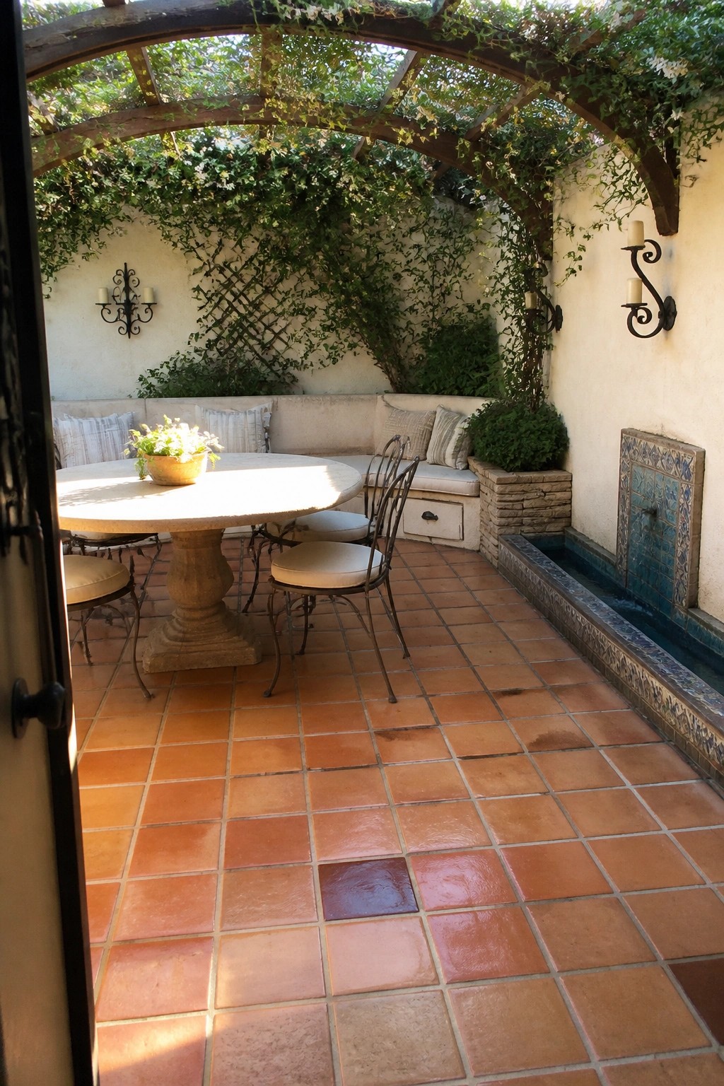 Sunlit terracotta patio with round table, vine-covered pergola, and tiled fountain.