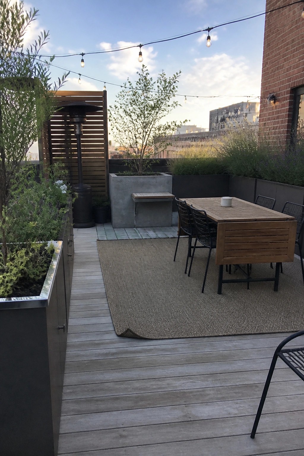 Rooftop deck with wooden dining table, chairs on rug, string lights, and planters at dusk