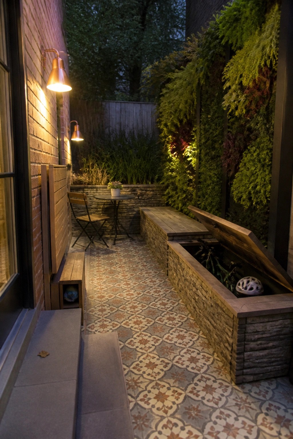 Narrow outdoor patio with patterned tile floor, stone and wood storage benches with one lid open, vertical garden wall, and wall mounted lights.