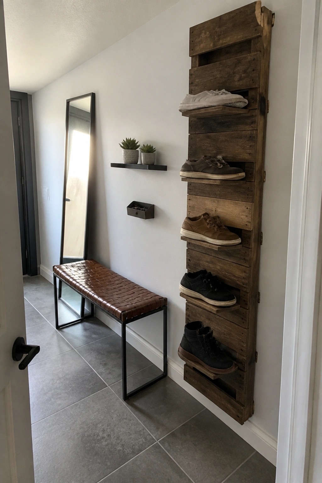 A vertical wooden pallet shoe rack mounted on a white wall in a narrow entryway holds several pairs of shoes, with a woven bench and mirror visible nearby.