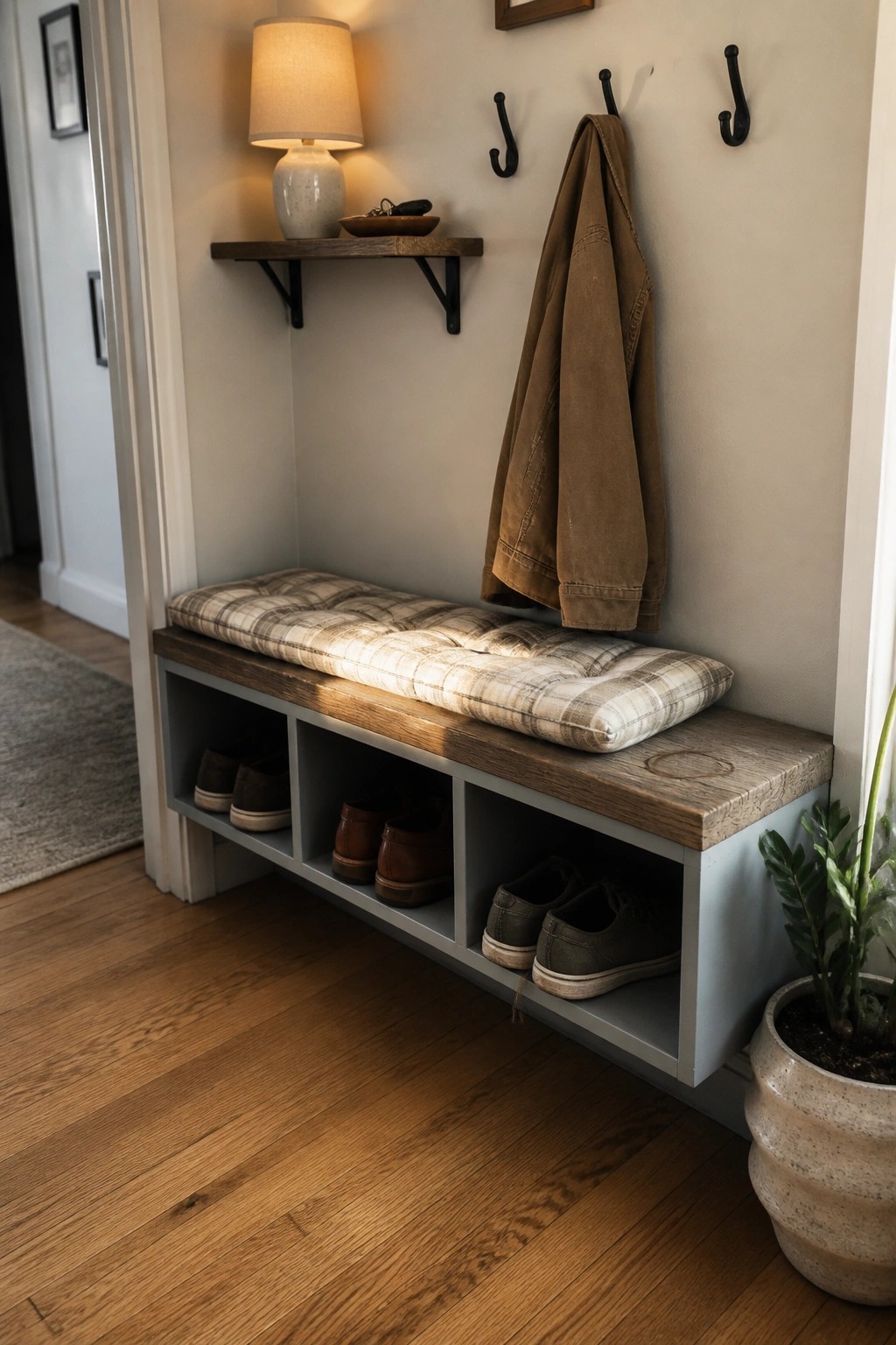 A wooden bench with gray-painted sides and open cubby compartments holding multiple pairs of shoes, topped with a plaid cushion in a narrow entryway.