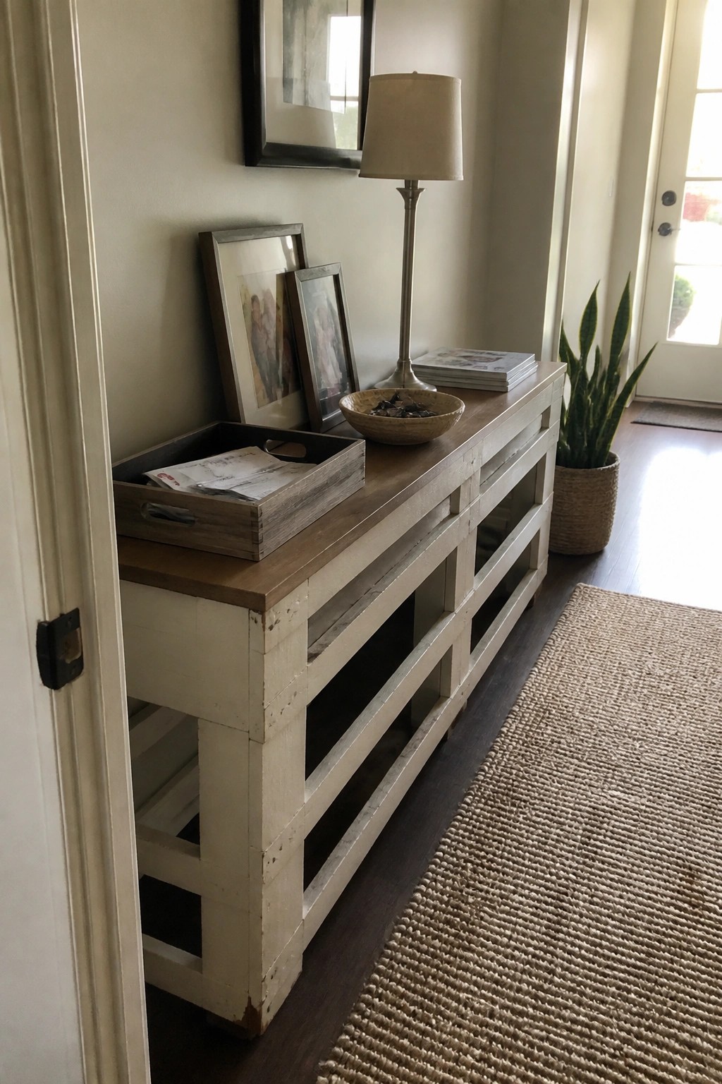 A whitewashed pallet console table with framed photos, a lamp, and a bowl sits against a wall in a hallway next to a woven rug.