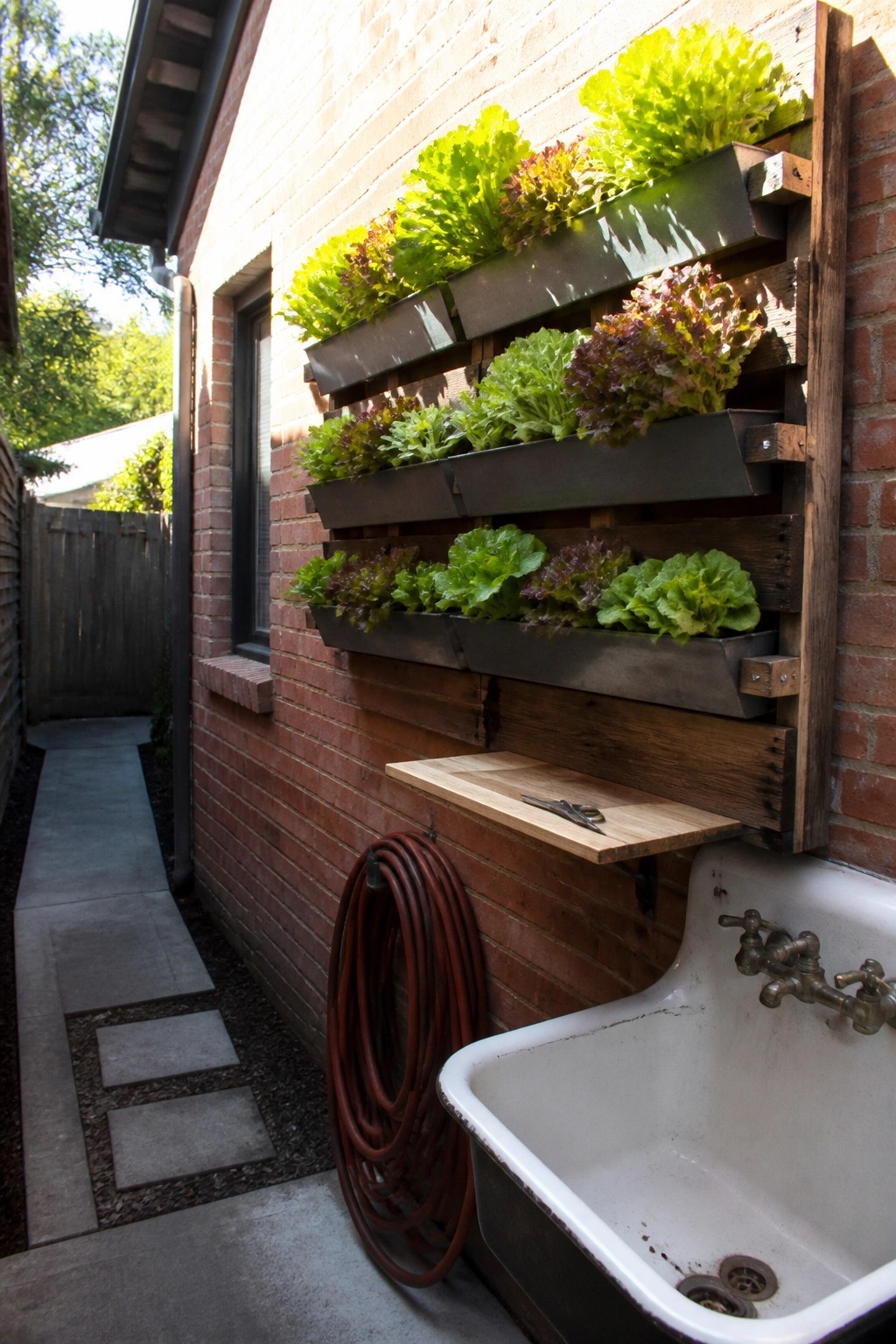 A vertical garden made from wooden pallets and metal troughs mounted on a brick wall, growing rows of green and red lettuce beside a white utility sink.