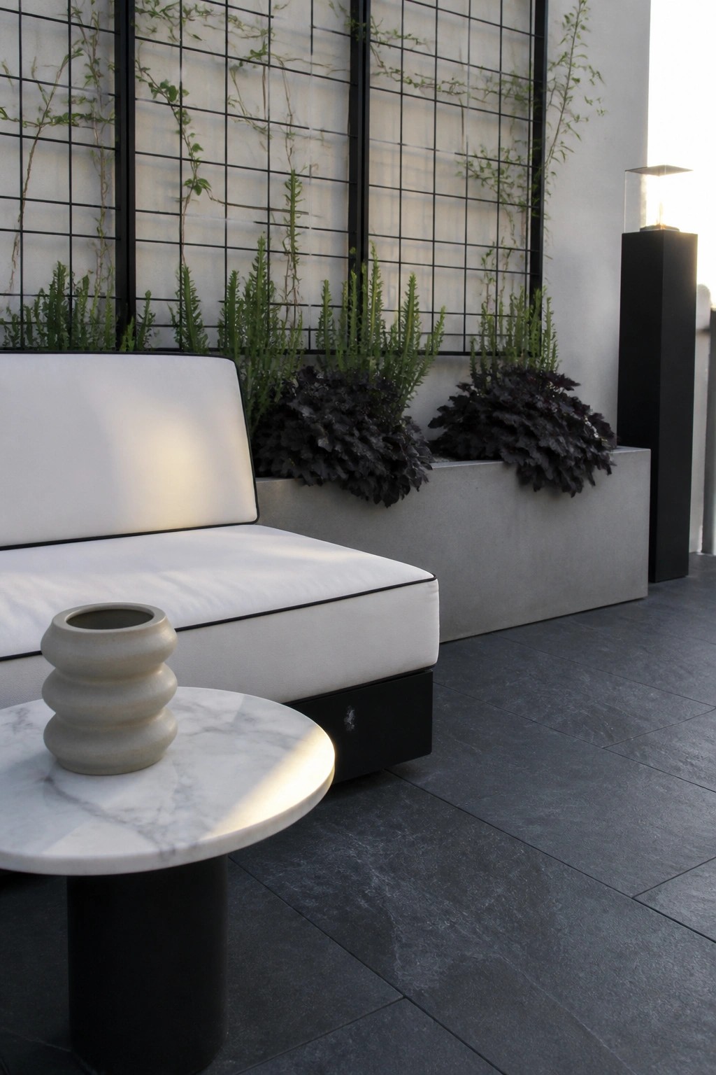 A white outdoor sofa sits beside a long concrete planter filled with green and dark foliage, with a black grid trellis behind it and a small marble table in the foreground.