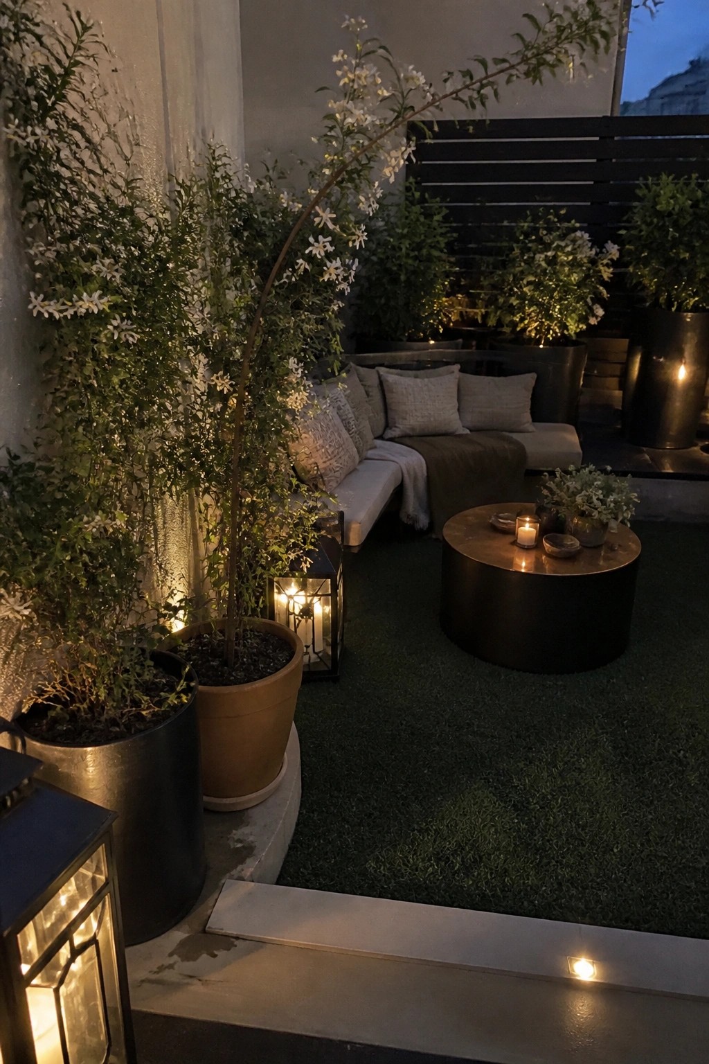 A nighttime patio scene with a curved outdoor sofa surrounded by large potted plants in metal and terracotta containers, a round coffee table with candles, and string lights on a dark fence.