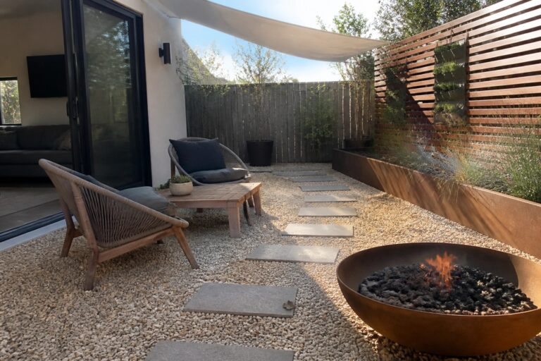 24 Affordable Backyard Patio Ideas Using Pea Gravel Backyard patio with pea gravel surface, stone paver path, large copper fire pit, two wicker chairs, and wooden coffee table beside a slatted fence with vertical garden.