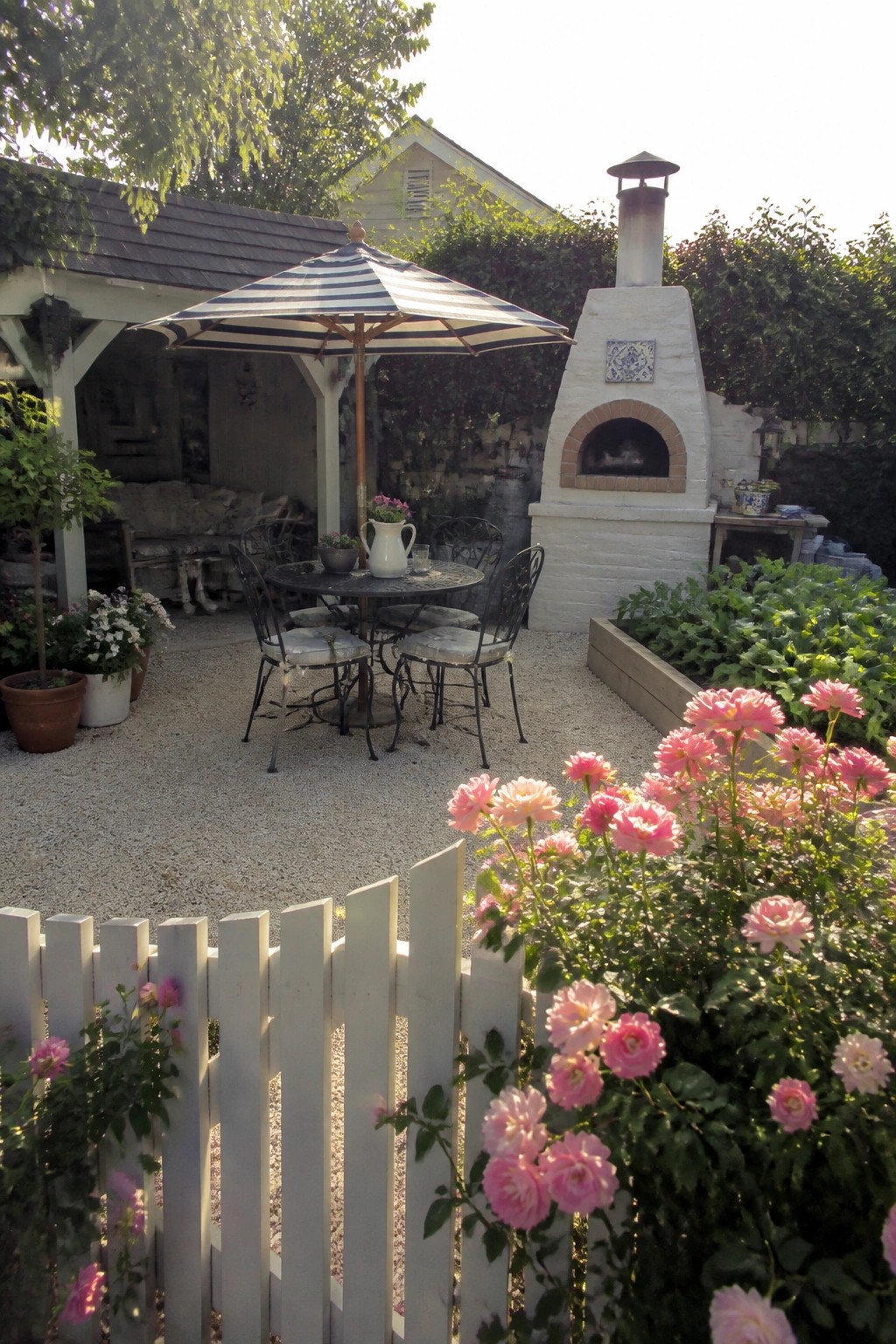 A backyard patio covered in pea gravel with a round metal table and chairs under a striped umbrella, positioned next to a white brick outdoor oven and bordered by pink roses.