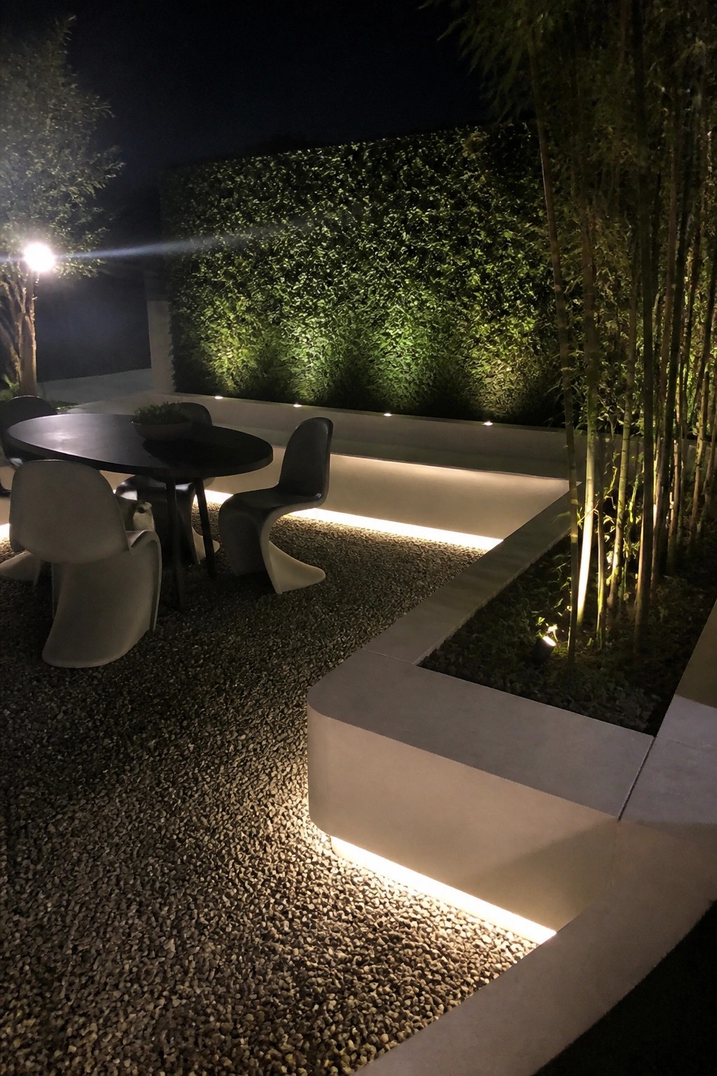 Nighttime outdoor patio with a round table and modern chairs on pea gravel, concrete borders, ground lighting, a green living wall, and tall bamboo plants.