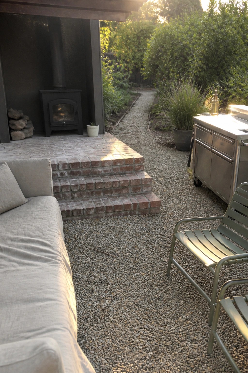 An outdoor space with pea gravel ground cover, a light gray sofa on the left, green metal chairs on the right, a stainless steel outdoor kitchen cart, brick steps leading to a black wood stove, and dense green shrubs in the background.