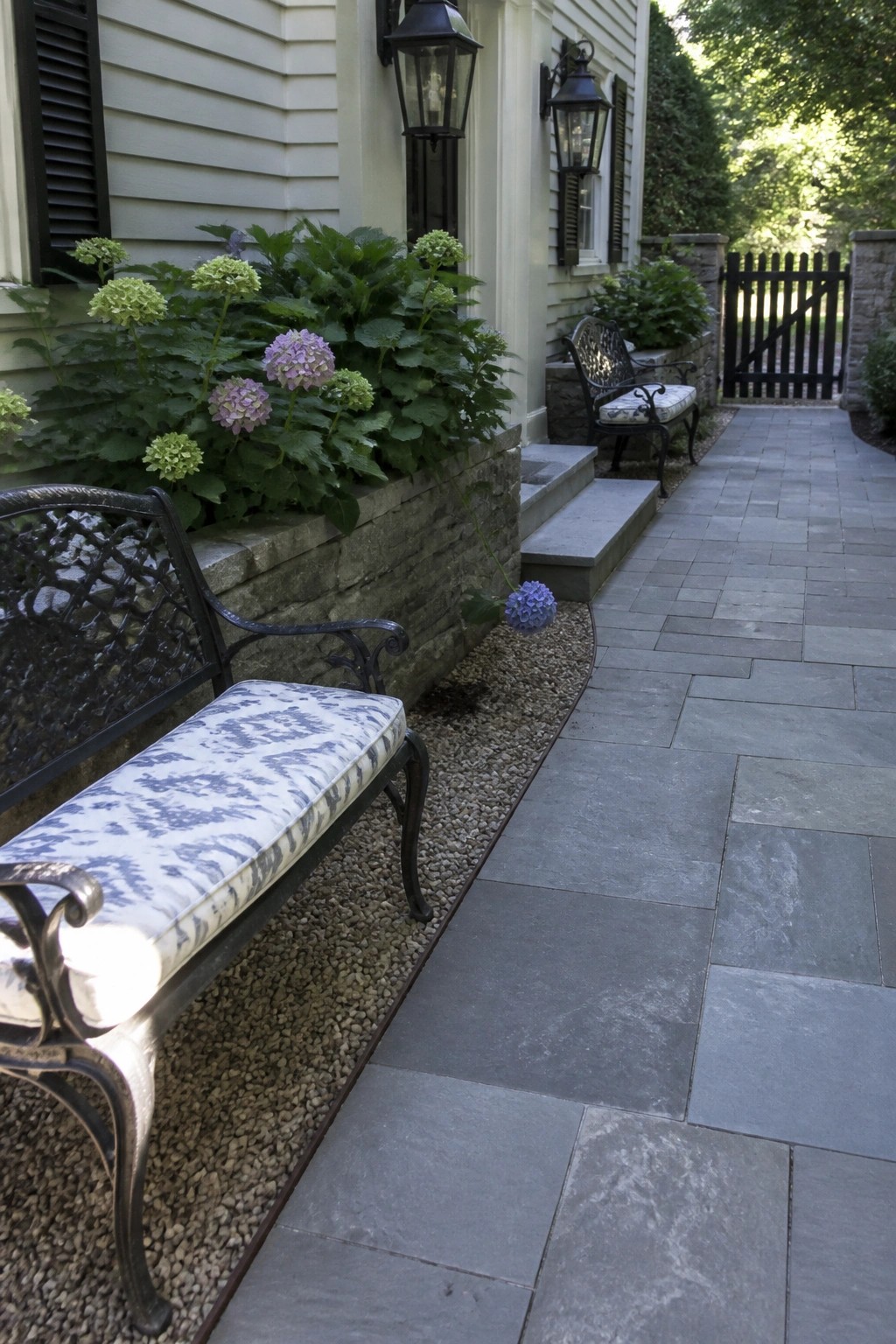 A gray stone paver walkway with a narrow gravel border running alongside a low stone retaining wall planted with hydrangeas.