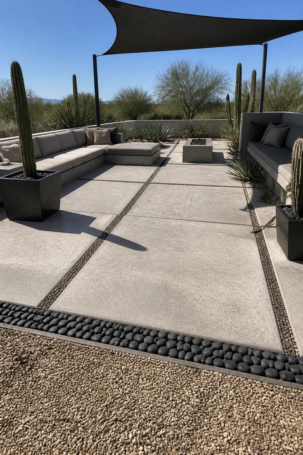 A concrete paver patio with dark pebble borders outlining rectangular sections, surrounded by built-in seating, potted cacti, and a fire pit under a shade sail.