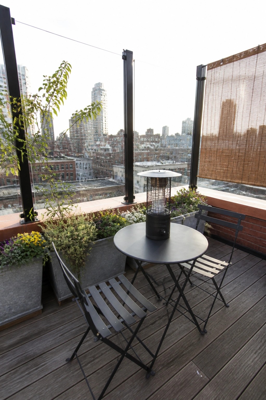 A small urban balcony with a round black dining table, two black folding chairs, a black patio heater, and several large planters filled with flowers and greenery overlooking city buildings.