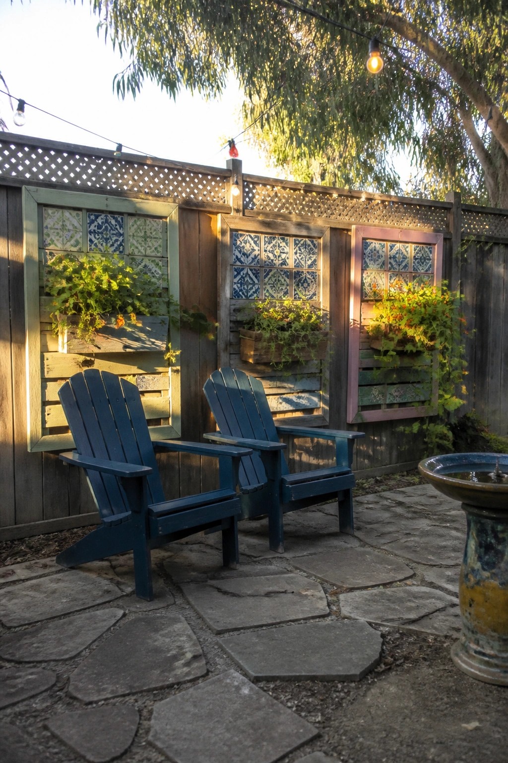 Two blue wooden chairs sit on a stone patio in front of a wooden fence holding three wall-mounted pallet planters filled with green plants.