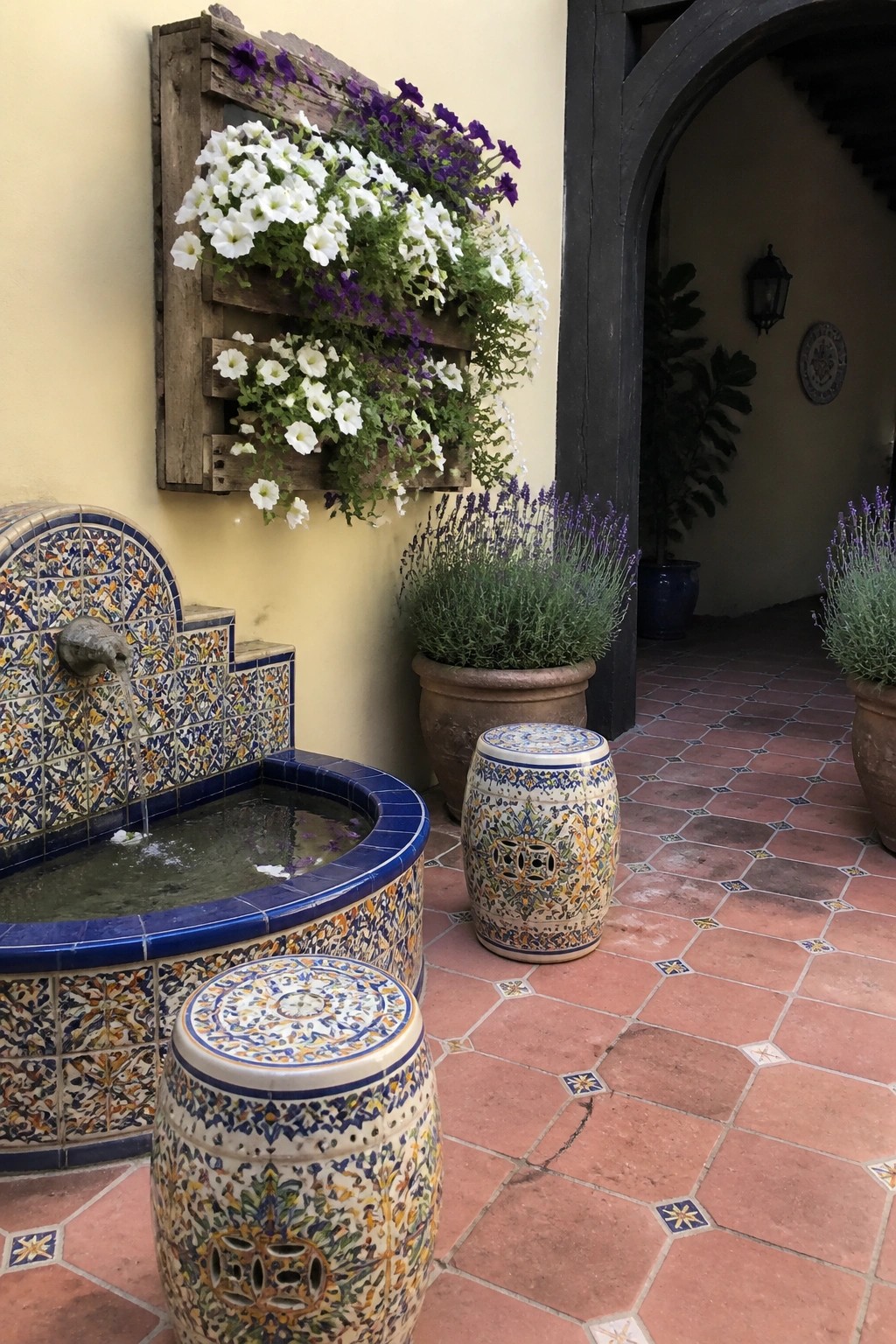 A wooden pallet mounted on a yellow exterior wall holds white and purple flowers above a tiled fountain on a terracotta patio.