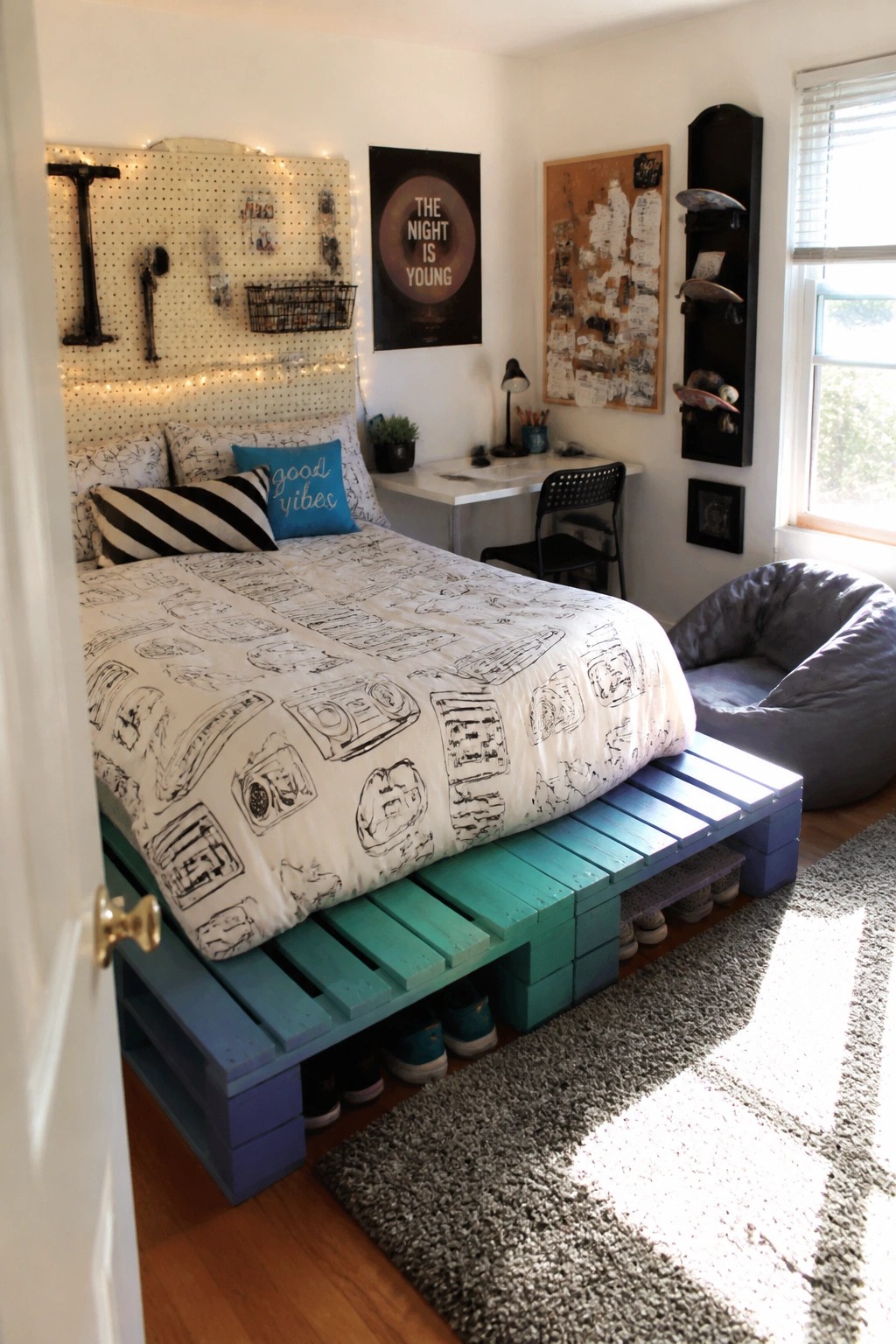 A bedroom with a bed on a low platform frame made of painted wooden pallets in blue and green tones, with patterned bedding and shoes stored underneath.