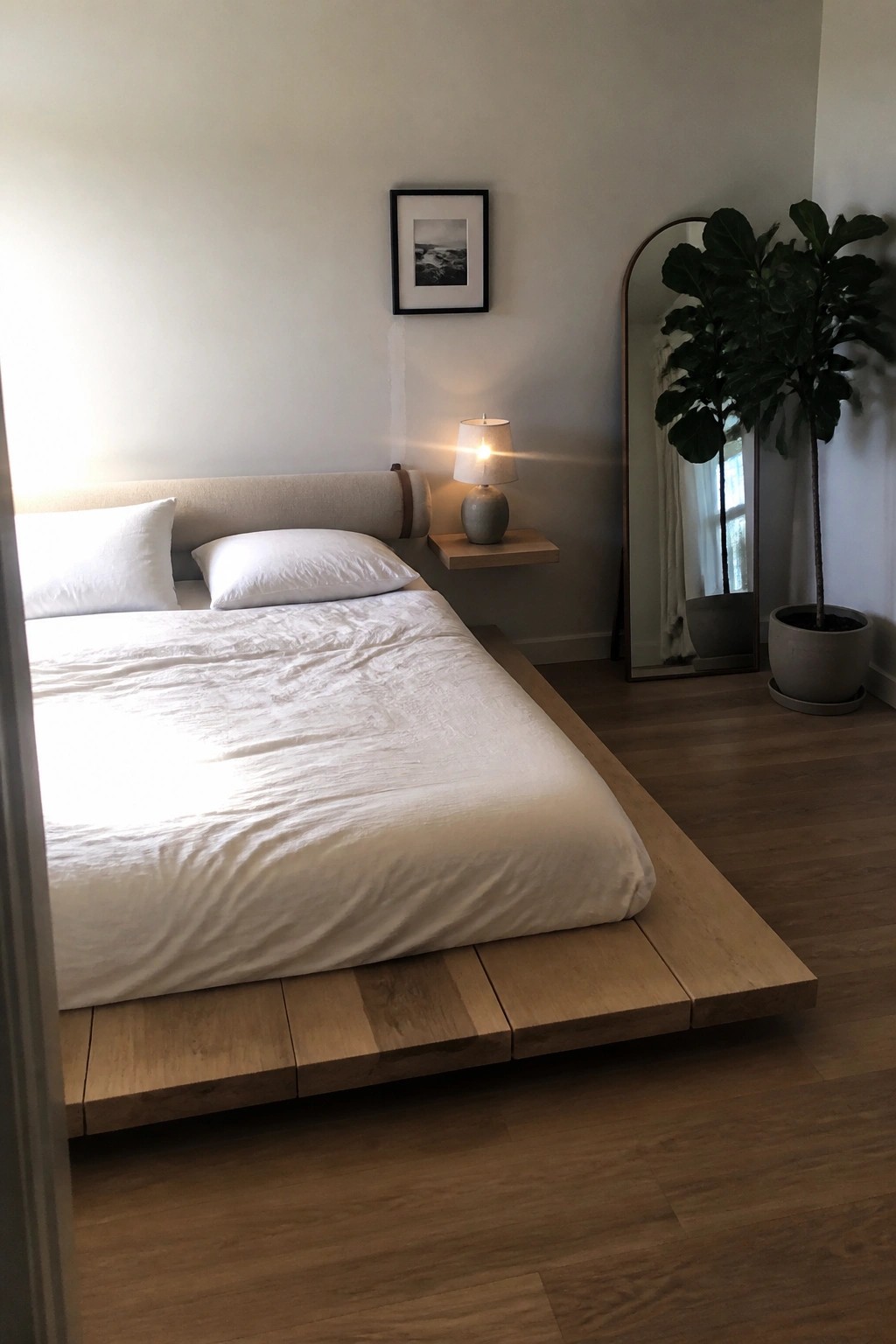 A bedroom with a low wooden platform bed covered in white linens, a floating nightstand holding a small lamp, and a tall arched mirror beside a potted plant.