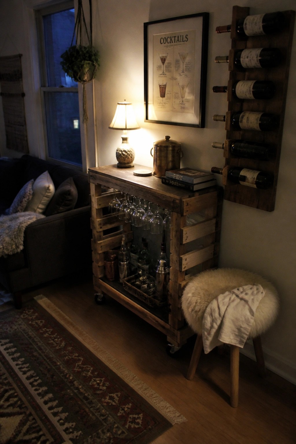 A rustic wooden pallet bar cart on wheels holds hanging wine glasses, bottles, and bar tools beneath a framed cocktail print, next to a wall-mounted pallet wine rack in a cozy living room.