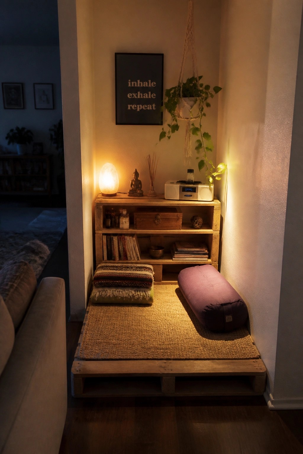 A small indoor corner with a wooden pallet platform covered by a woven rug, holding stacked cushions and a purple bolster beside a low shelf with books and a lit lamp.