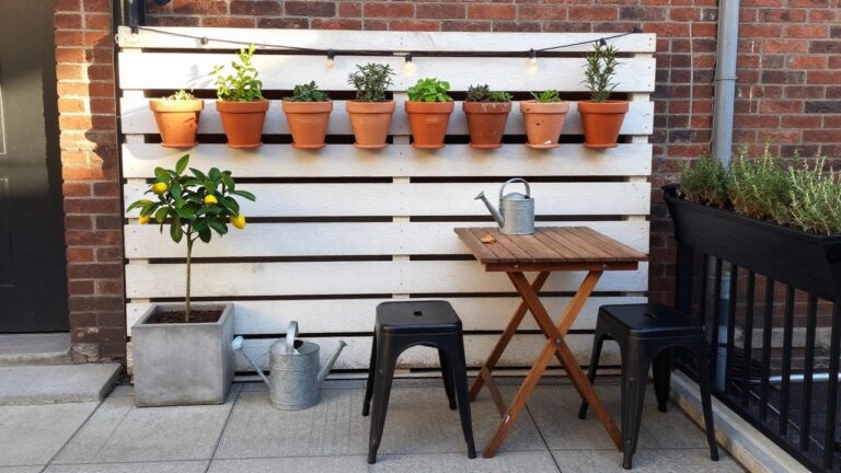 22 Smart Vertical Pallet Garden Ideas for Patios With Limited Floor Space A compact outdoor patio with white-painted wooden pallets mounted on brick walls holding rows of terracotta plant pots, a small wooden table, and two black stools.