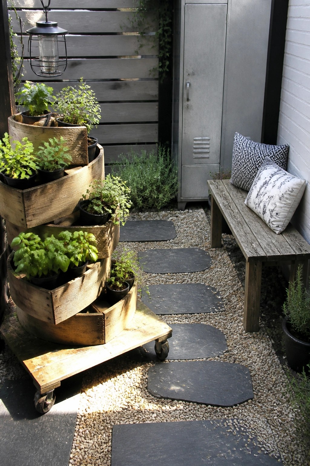 Mobile wooden crate planters with herbs beside bench with pillows on gravel path