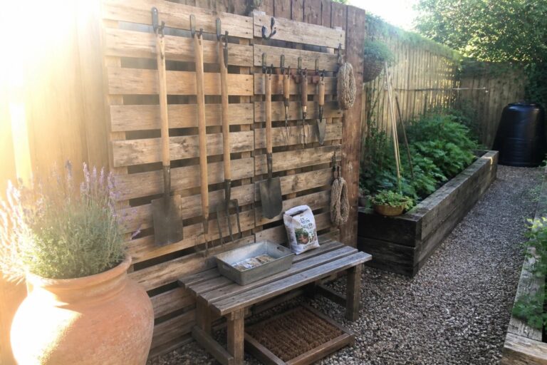 25 Useful Outdoor Pallet Storage Ideas for Long-Handled Garden Tools A weathered wooden pallet stands upright against a fence with garden tools hanging from metal hooks and a wooden bench holding boots and a metal tray below.