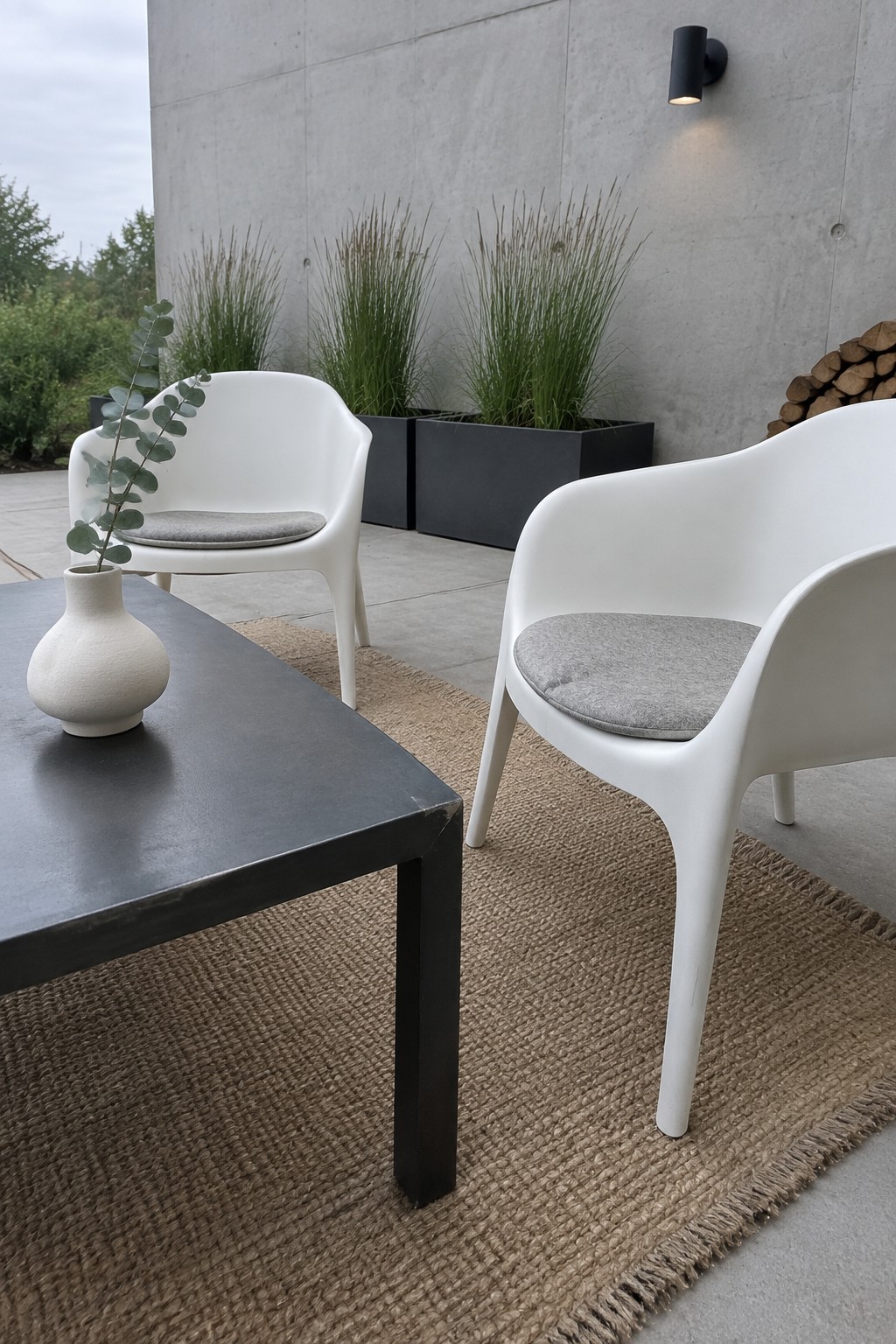 White molded chairs with gray cushions sit around a dark patio table on a large woven jute rug over concrete, with planters and a wall light in the background.