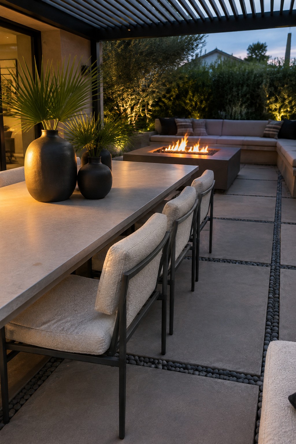 A long concrete outdoor dining table with upholstered chairs holds two large dark ceramic vases with tall green plants, set on a stone patio near a fire pit lounge area.