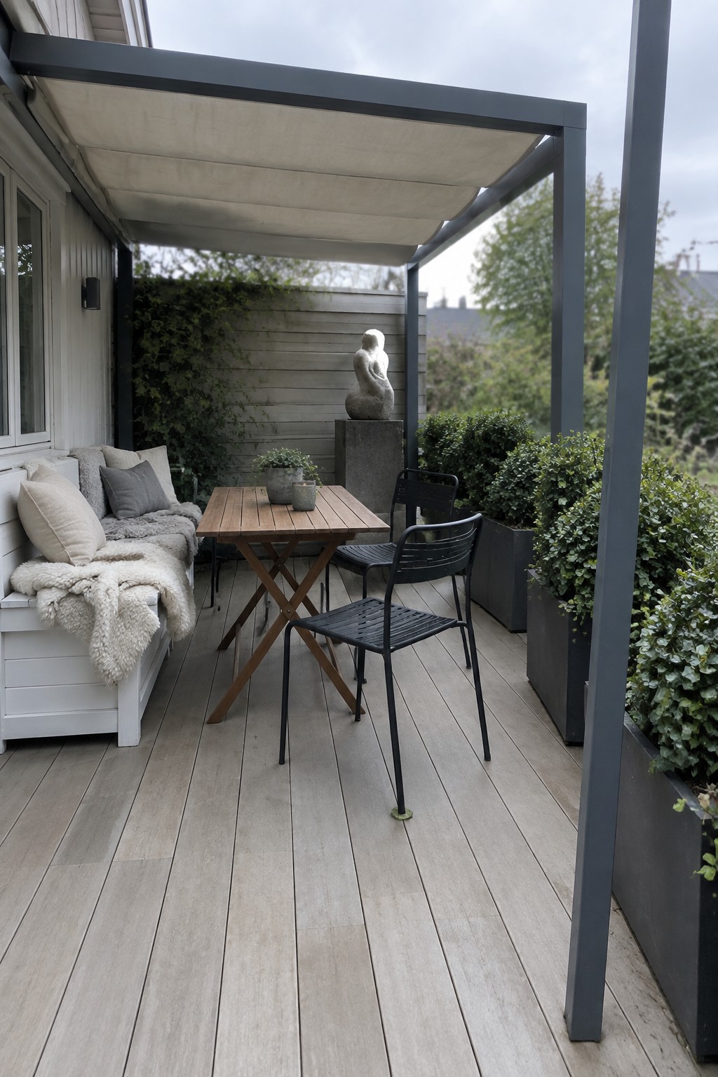A small wooden deck patio with a gray metal pergola, white cushioned bench, wooden folding table, black metal chairs, and large planters.