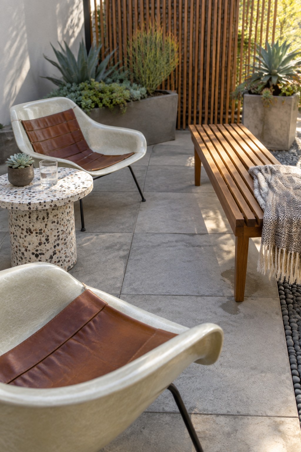 An outdoor patio with two modern lounge chairs, a round terrazzo side table, a long wooden bench, potted plants, and a tall vertical wooden slat screen in the background.