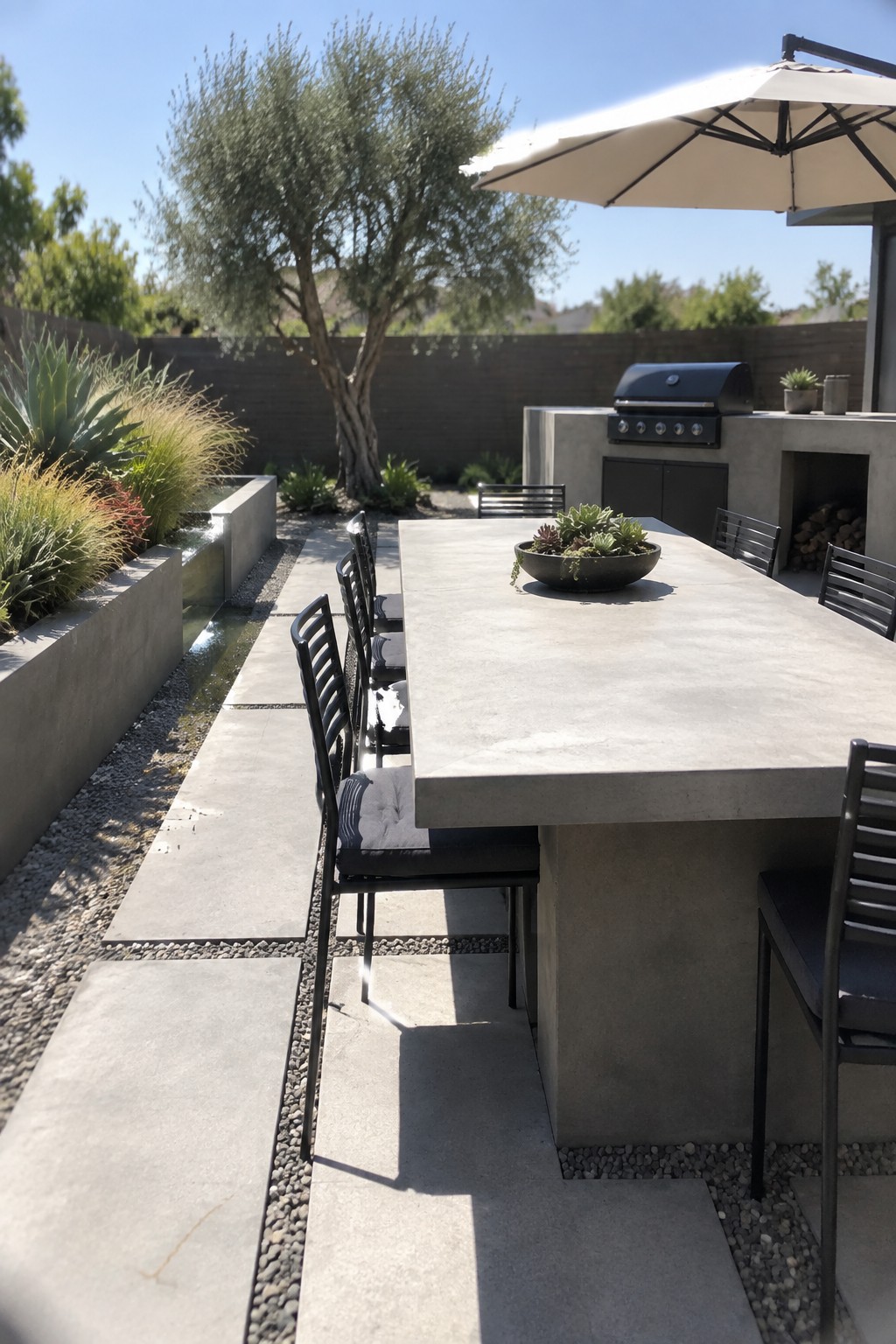 Modern concrete dining table with black chairs on sunny backyard patio with water feature