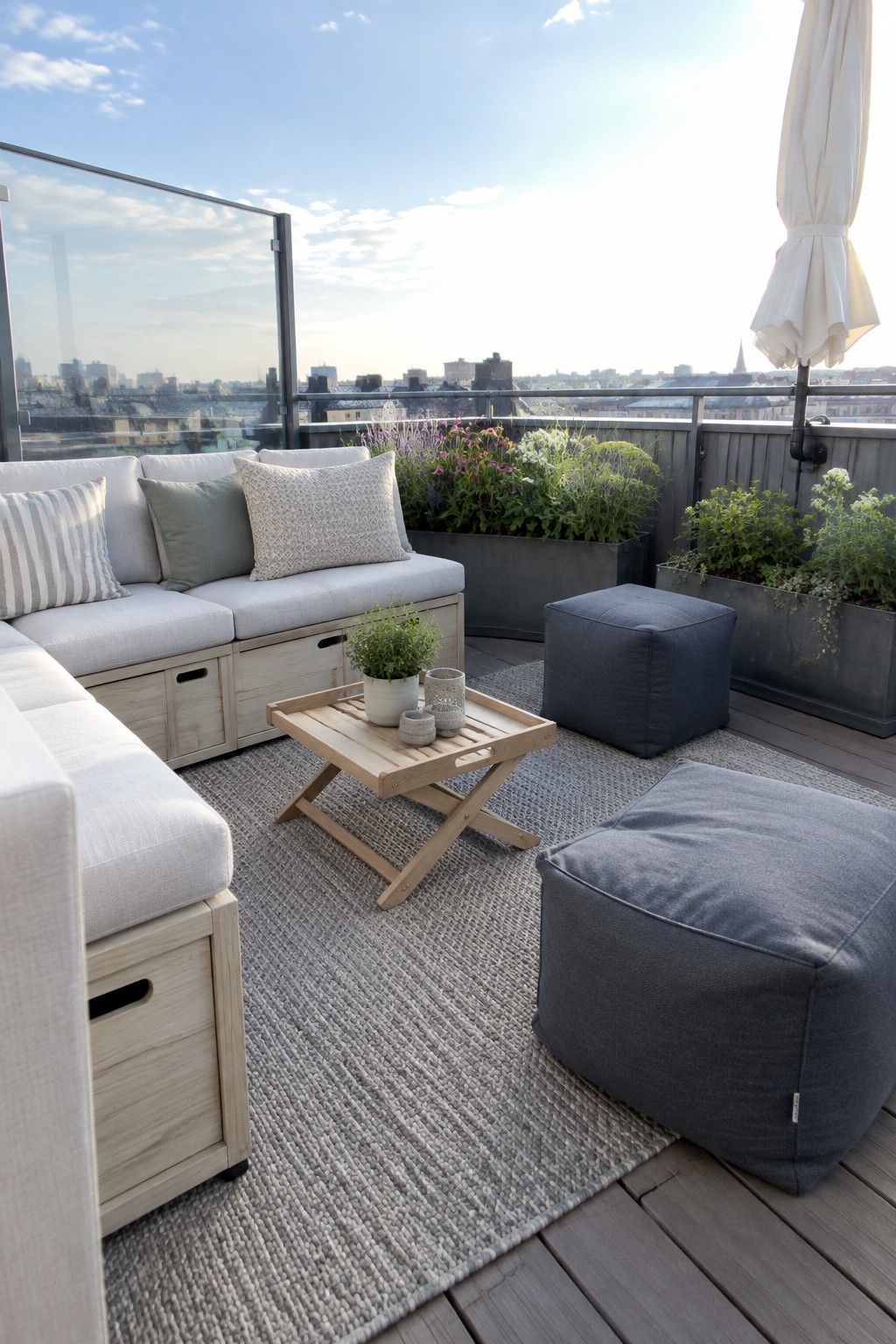 A rooftop patio featuring a light wood sectional sofa with storage, two gray fabric poufs, a wooden coffee table, and large planters on a woven rug with a city view in the background.