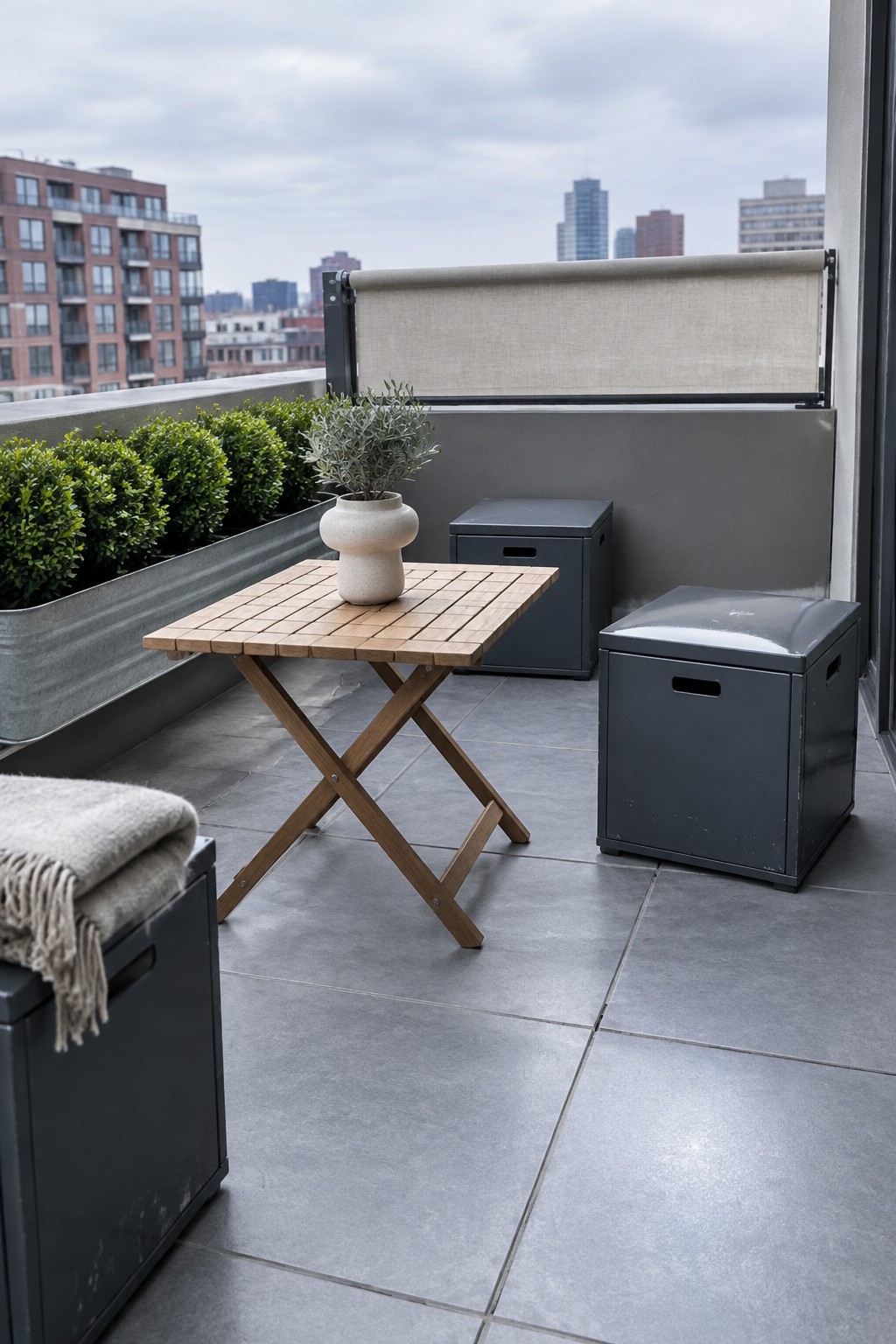 A small apartment balcony with a wooden folding table, two dark gray storage boxes, potted plants in a metal planter, and a beige shade screen overlooking city buildings.