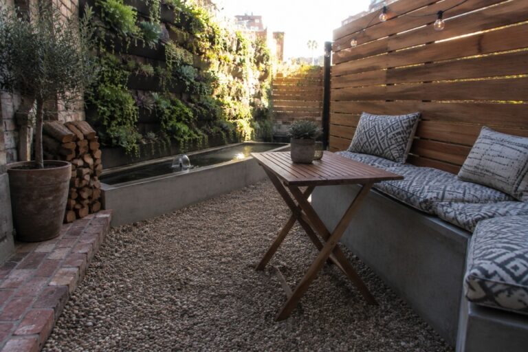 18 Small Patio Retreat Ideas That Create a Private, Comfortable Outdoor Escape A small gravel patio with a concrete bench, patterned cushions, wooden folding table, and horizontal wood slat privacy screen beside a living wall and water feature.