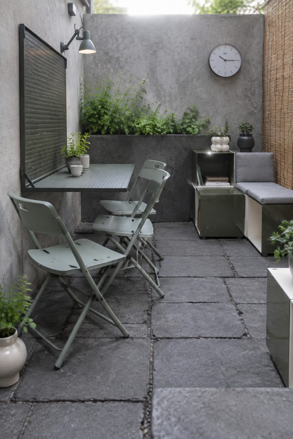 A compact outdoor patio with stone pavers shows several sage green foldable metal chairs, a matching table, a storage bench with gray cushions, and potted plants against concrete walls.