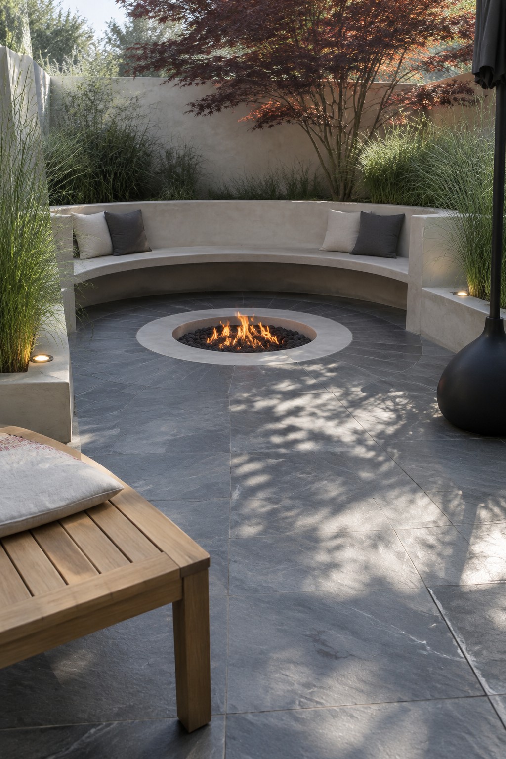 A round fire pit set in dark stone tiles is surrounded by a curved built-in concrete bench with cushions, bordered by tall grasses and a red-leafed tree.