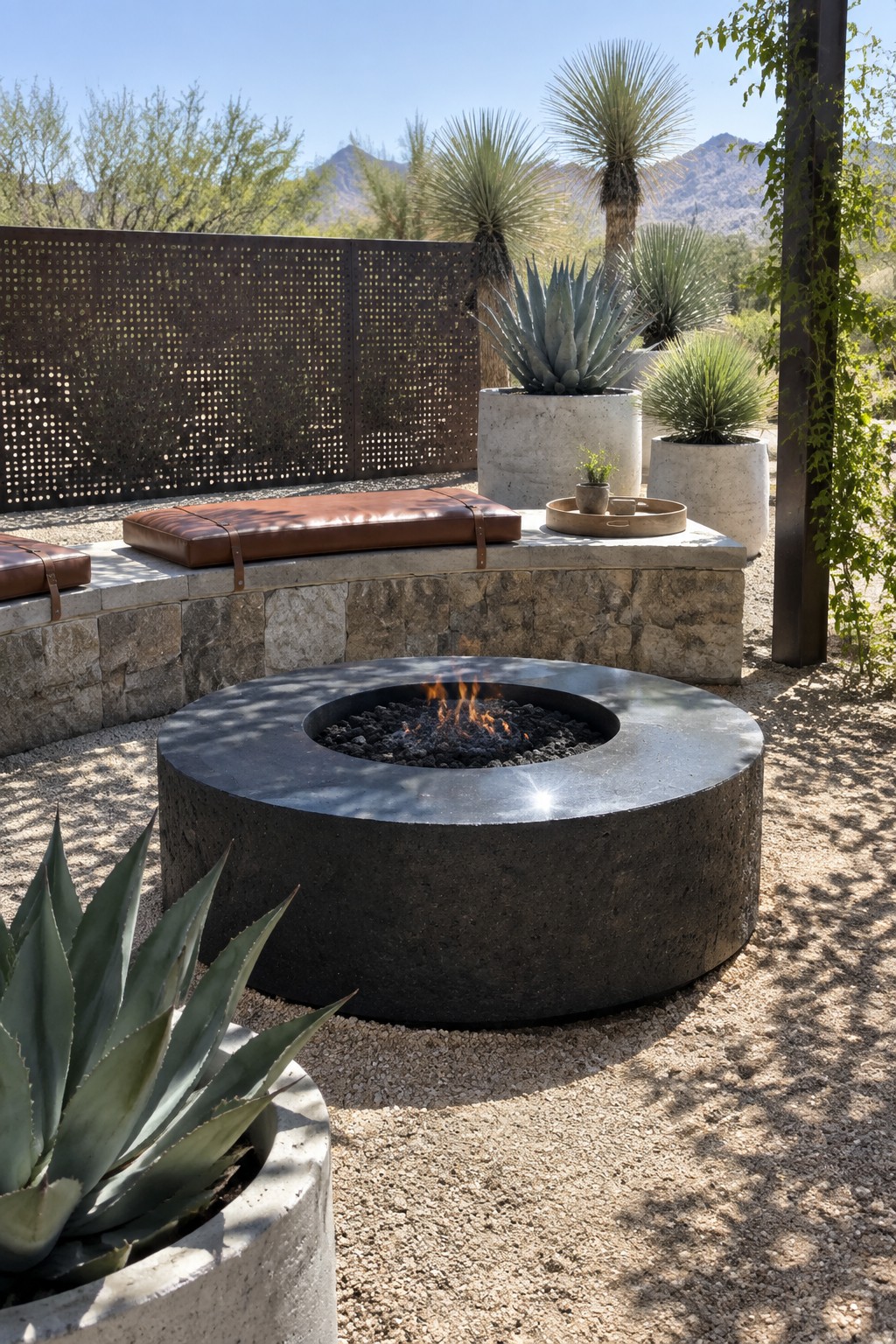 A round dark concrete fire pit with a burning fire sits on gravel in front of a curved stone bench holding brown leather cushions, with large potted desert plants and a perforated metal screen in the background.