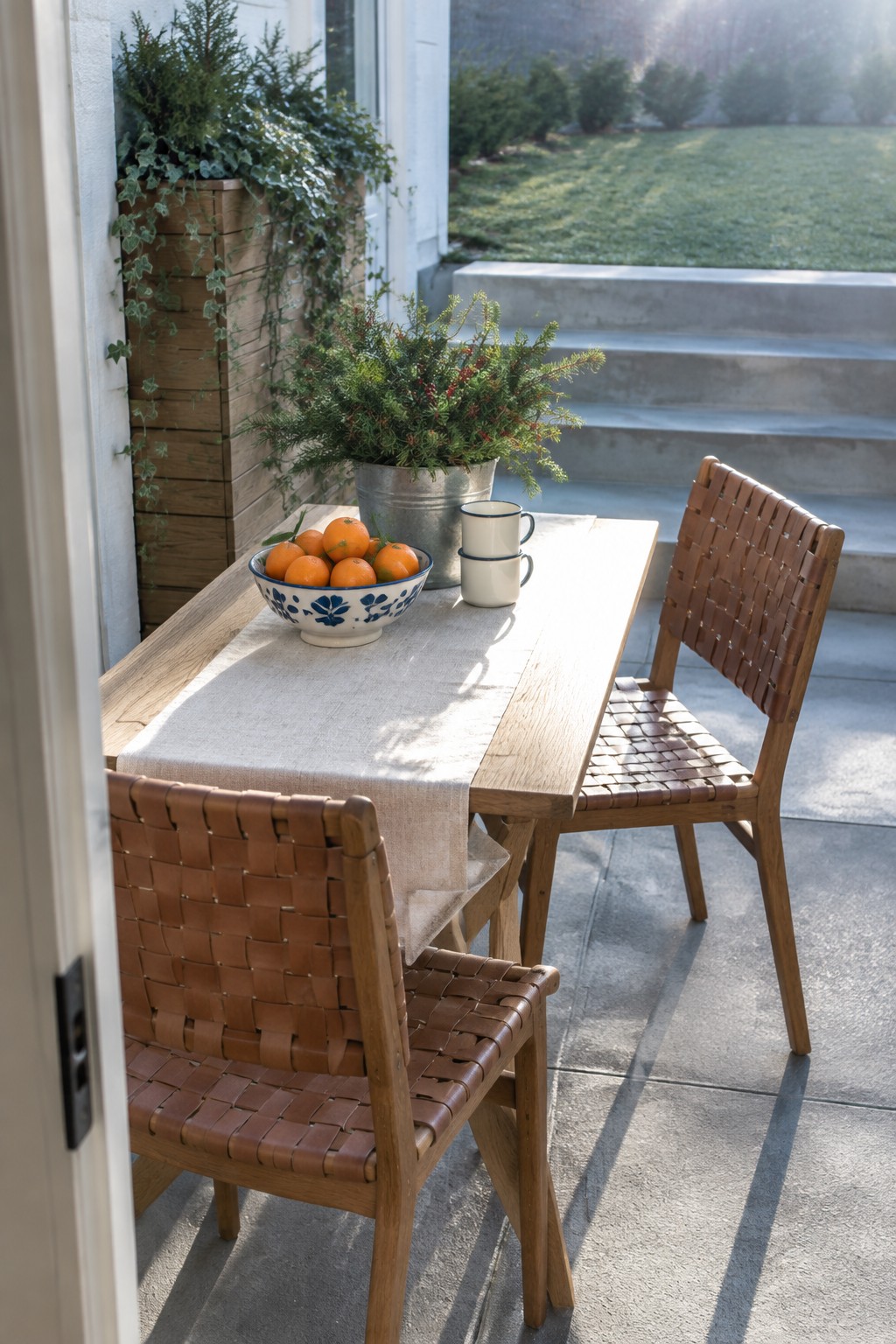 A wooden patio table on a concrete surface holds a linen runner, a blue and white bowl of oranges, two stacked white mugs, and a metal bucket with evergreen branches and red berries, with two woven leather chairs pulled up to the table.