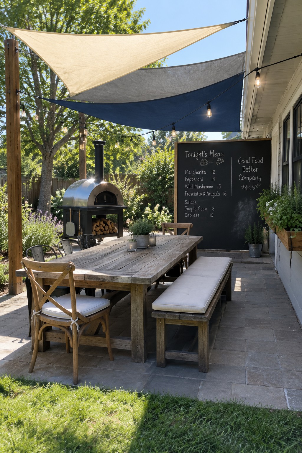 A stone patio with a wooden dining table and bench shaded by three overlapping triangular sail shades in beige, gray, and blue, next to a pizza oven and chalkboard menu.