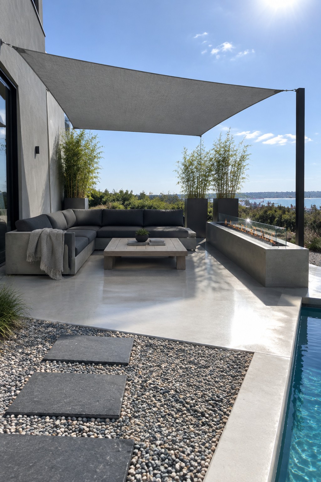 A modern patio with a large gray triangular sail shade stretched over a dark sectional sofa, a low wooden coffee table, a long concrete fire pit with flames, and a swimming pool on the right side.