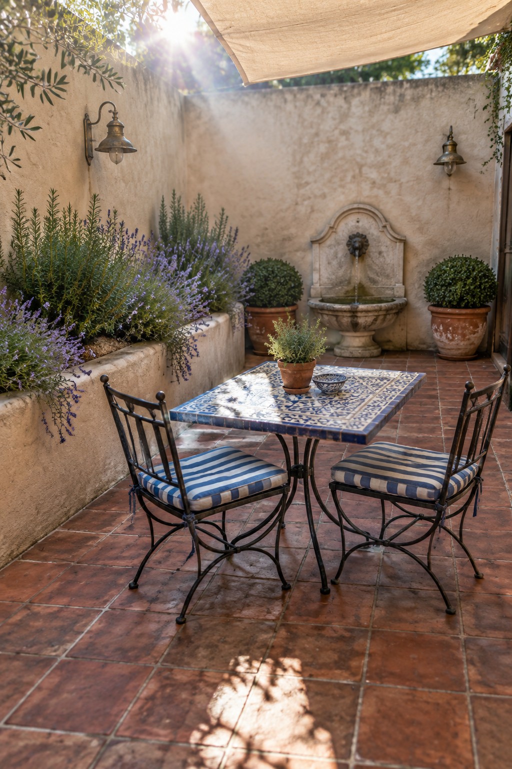 A small outdoor patio with a blue and white tiled square table, two black metal chairs with blue and white striped cushions, a stone fountain on the back wall, and potted plants along stucco walls on terracotta tile flooring.
